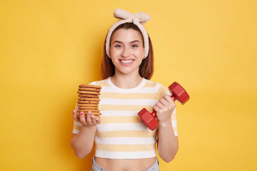 Extremely happy young dark haired woman wearing striped shirt and hair band holding red dumbbell and cookies, posing isolated over yellow background, looking at camera with toothy smile.